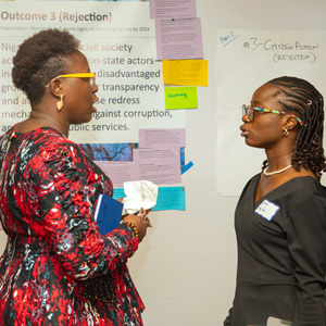 Two individuals conversing in front of a wall with assorted notes and papers.